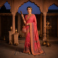 Woman in traditional pink saree with gold patterns standing in an ornate indoor setting.